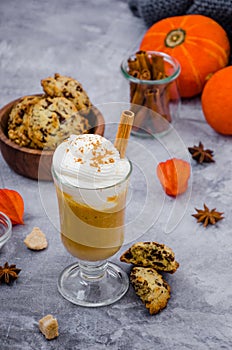 Pumpkin latte in a glass with spicy cream and a cinnamon stick on a gray stone background with cookies with chocolate chips.