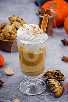 Pumpkin latte in a glass with spicy cream and a cinnamon stick on a gray stone background with cookies with chocolate chips.