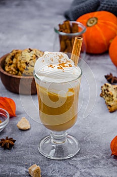 Pumpkin latte in a glass with spicy cream and a cinnamon stick on a gray stone background with cookies with chocolate chips.