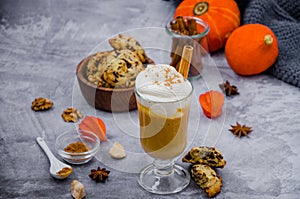 Pumpkin latte in a glass with spicy cream and a cinnamon stick on a gray stone background with cookies with chocolate chips.