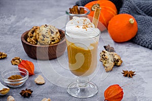 Pumpkin latte in a glass with spicy cream and a cinnamon stick on a gray stone background with cookies with chocolate chips.
