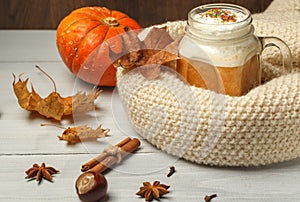 Pumpkin latte with thick milk foam and spices, in a glass mug, wrapped in a knitted white scarf. On a light wooden table with