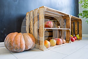 Pumpkin next to wooden box filled with fruit and preservation on floor in modern kitchen on background of black wall. Autumn harve