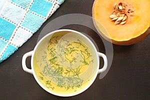Pumpkin puree soup is in a round plate with two handles. Nearby there is a fresh pumpkin with seeds and a kitchen towel