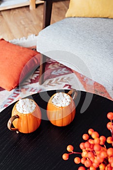Pumpkin shaped cups of hot drink with marshmallows on black wooden table in cozy interior. Pumpkin latte. Cozy autumn