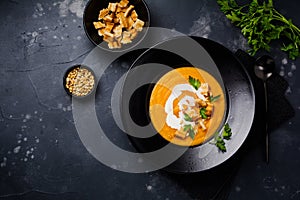 Pumpkin soup with cream, pieces of bread and cedar nuts in black ceramic plate on dark wooden background.