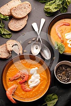 Pumpkin soup with shrimps in two dark bowls next to bread, seeds and silver spoons