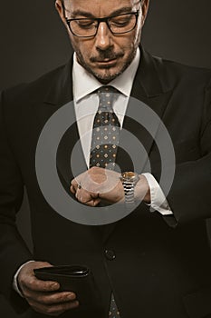 Punctual businessman checks time looking on his wristwatch while holding wallet in other hand. Well dressed bearded man