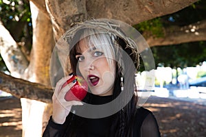 Punky girl with blonde and brown hair holds a red apple in her hands to bite it. the girl wears a crown of thorns like christ on