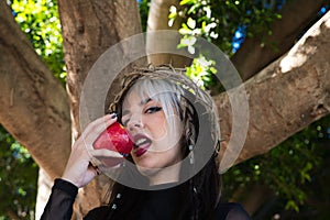 Punky girl with blonde and brown hair holds a red apple in her hands to bite it. the girl wears a crown of thorns like christ on