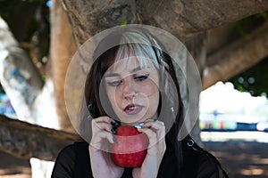 Punky girl with blonde and brunette hair holds a red apple in her hands and stares at it. The girl is a witch from the fairy tale