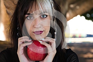 Punky girl with blonde and brunette hair holds a red apple in her hands and stares at it. The girl is a witch from the fairy tale