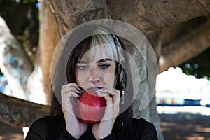 Punky girl with blonde and brunette hair holds a red apple in her hands and stares at it. The girl is a witch from the fairy tale