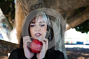 Punky girl with blonde and brunette hair holds a red apple in her hands and stares at it. The girl is a witch from the fairy tale