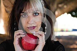 Punky girl with blonde and brunette hair holds a red apple in her hands and stares at it. The girl is a witch from the fairy tale