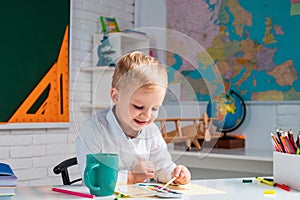 Pupil child is sitting at a desk indoors. Cute pupil with funny face schooling work. Kids gets ready for school.