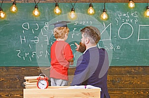 Pupil in graduation cap solving equation with the help of the teacher. Man teaching little kid arithmetic while pointing