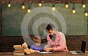 Pupil with teacher at school. Tutor helping kid to write letters in copybook. Man and boy sit at desk with laptop and