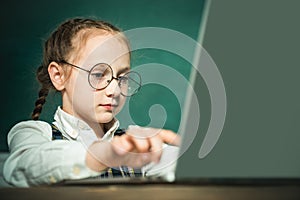 Pupil working on laptop computer over blackboard background. Child near chalkboard in school classroom. Kid is learning