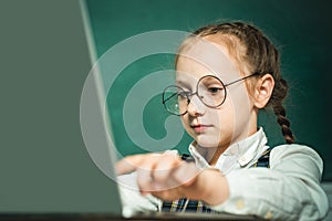 Pupil working on laptop computer over blackboard background. Child near chalkboard in school classroom. Kid is learning