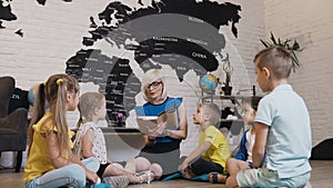 Pupils sit in circle on floor on the pillows as teacher reads to them interesting book in class at elementary school or