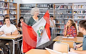 Pupils sitting in class and listening carefully to female teacher holding Peru flag in hands and talking about geography