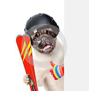 Puppy in helmet holding skis, peeking from behind empty board and showing thumbs up. isolated on white background