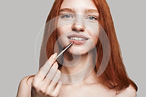 Pure beauty. Portrait of cute young woman with long red hair painting lips with lipliner and looking at camera while