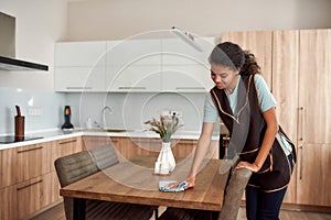 Pure clean. Young positive afro american house maid in apron cleaning table in the modern kitchen. Professional female