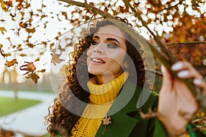 Pure gentle look and white-toothed smile at the curly brunette holding her hand on a thin branch of autumn tree in the