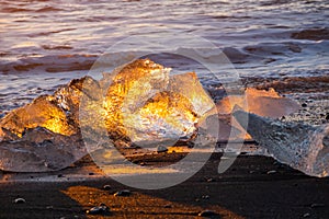 Pure Ice Shining on Black Volcanic Sand at Sunset. Icebergs from Glacier in Sun Rays. Clear Ice in Ocean Waves. Famous