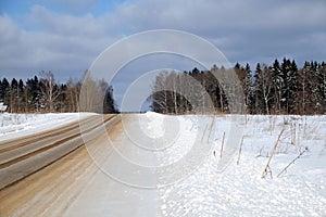 Purified empty suburban sand-strewn road through a snowfield and a forest under blue sky with white clouds on frosty bright sunny