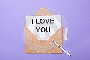 On a purple background, a white pen, a craft envelope and a white card with the text I LOVE YOU. View from above