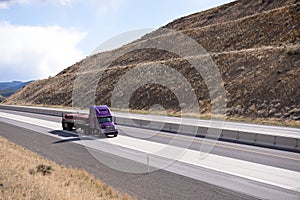 Purple big rig semi truck with flat bed semi trailer transporting covered cargo on the road between the hills in Oregon