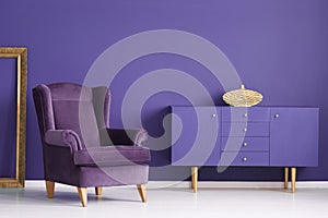 Purple cabinet with a golden vase, comfy armchair and frame in a