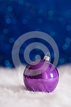 Purple Christmas ball on white fur with garland lights on blue b