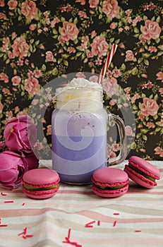 Purple coffee in stylized mason jar cup with macarons and roses and colorful decoration on background of floral pattern. Blueberry