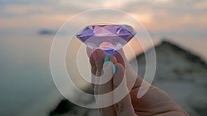 Purple diamond crystal in a female hand, close up, on sunset background