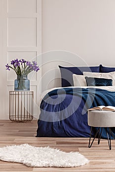 Purple flowers in blue glass vase on stylish bedside table next to king size bed