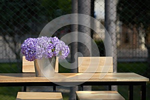 Purple flowers in bucket vase on wooden table with chairs with s