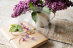 Purple lilac bouquet in vase and present laid on wooden table.