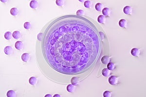 Purple marbles in drinking transparent glass with scattered marbles on white background top view