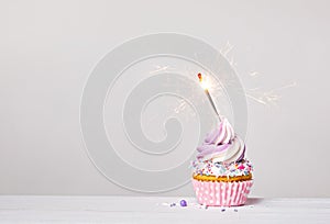 Purple and pink cupcake with sprinkles and lit sparkler on a white grey background.