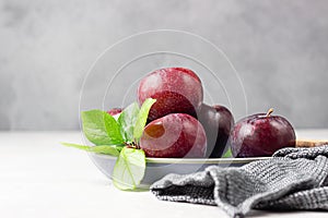 Purple plums with leaves and knife on ceramic plate, light grey concrete background