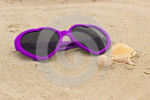 Purple sunglasses shaped heart with shells on the sand