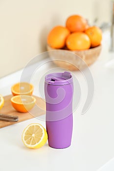 Purple thermo mug with freshly squeezed orange juice on the background of a cup with oranges. Healthy food concept.
