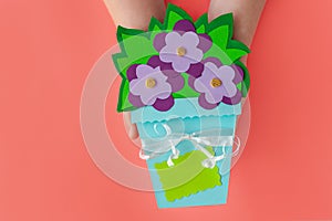 Purple violet paper flowers with green leaves in basket on pink coral background with greeting card tag mockup flat lay