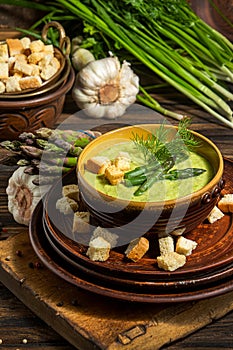 Purred creamy asparagus soup in glass bowl on black plate against raw fresh asparagus and greenery