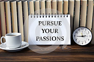 PURSUE YOUR PASSIONS - words on white notebook on background of books, clock and cup of coffee