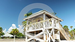 Push out footage of a white wooden lifeguard tower on a sandy white beach with lush green palm trees and blue ocean water